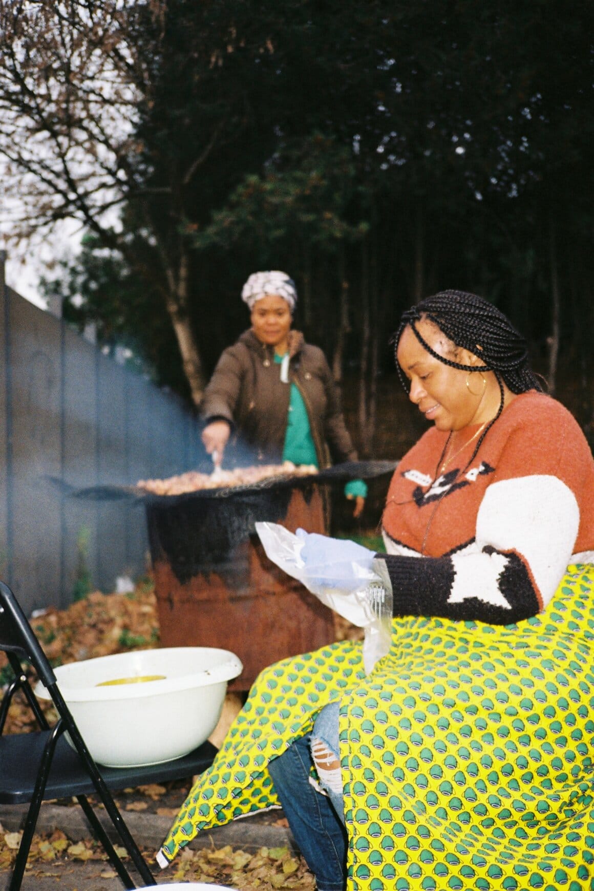 Une femme manipule un sac en plastique tandis qu’une autre prépare à manger sur un barbecue à l’arrière-plan.