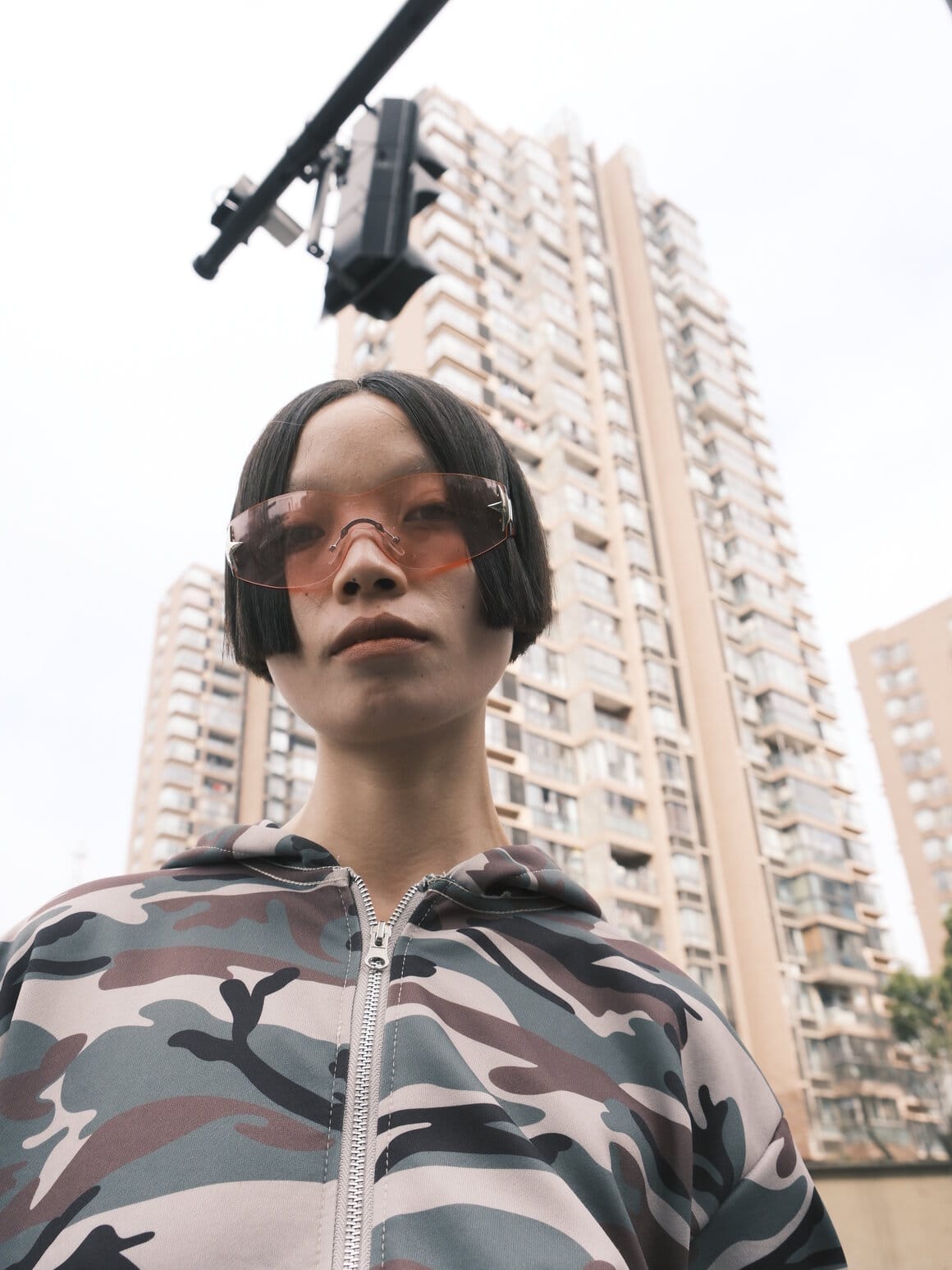 Une femme portant de grandes lunettes teintées pose en contre-plongée devant plusieurs tours d’habitation.