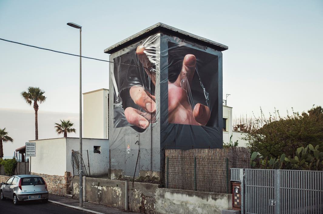 Fresque murale réalisé par l'artiste Shozy. La fresque imite les autres murs de l'immeuble mais modifie le relief en donnant l'illusion qu'une main est à l'intérieur du batiment et tente d'en sortir en déchirant le film plastique qui enveloppe la structure